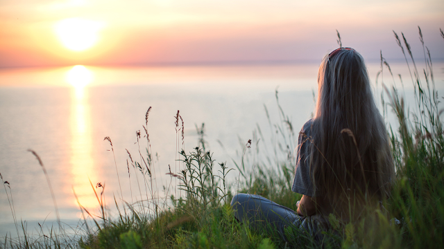 Pessoa sentada na relva a contemplar o pôr do sol sobre o mar, num momento de tranquilidade e reflexão. Reflexão ao pôr do sol – Dia Mundial da Paz e do Desenvolvimento