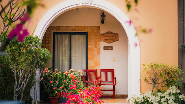 Inspiração: Um Refúgio Acolhedor no seu Espaço Exterior Entrada de uma casa com um arco branco sobre uma porta de vidro, decorada com duas cadeiras de madeira pintadas de vermelho. O ambiente é rodeado por plantas exuberantes com flores vermelhas e brancas, criando uma atmosfera de tranquilidade e um espaço exterior convidativo.