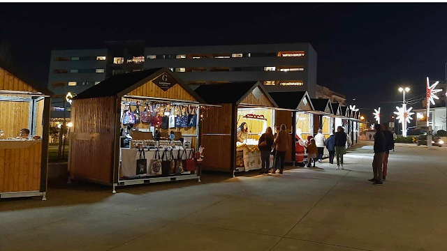 Mercado de Natal: O Charme das Casinhas de Madeira ao Anoitecer Fotografia noturna de um mercado de Natal ao ar livre. Em primeiro plano, vê-se uma fila de pequenas cabanas de madeira iluminadas, onde artesãos expõem produtos como malas e decorações. Várias pessoas passeiam calmamente pelo recinto pavimentado. Ao fundo, luzes de Natal em forma de estrela decoram os postes de iluminação e um edifício moderno destaca-se no horizonte sob o céu escuro.