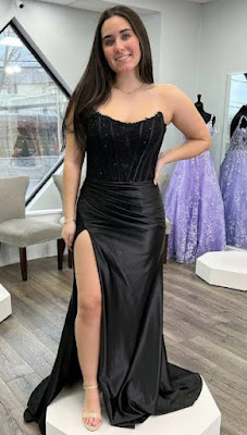 Vestido de gala preto com corpete estruturado e saia de cetim com fenda lateral Uma mulher jovem de cabelos longos e escuros sorri enquanto posa com um vestido de festa preto comprido. O vestido destaca-se pelo contraste de materiais: o topo é um corpete justo, sem alças (tomara-que-caia), com detalhes bordados e transparências subtis, enquanto a saia é feita de um cetim preto brilhante, apresentando um drapeado na anca e uma fenda alta na perna direita. Ela está em cima de uma plataforma branca num salão de festas; ao fundo, veem-se poltronas cinzentas, uma janela ampla e outros vestidos de gala em tons de lilás expostos em manequins. O chão é de madeira clara.