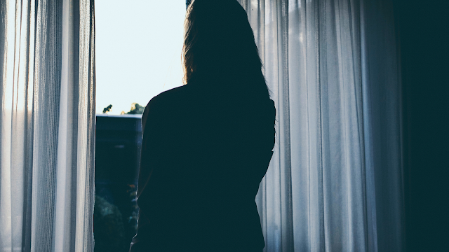 A forma como enfrentamos o sofrimento Pessoa de pé junto a uma janela, olhando para o exterior através das cortinas, num momento de reflexão e introspeção.