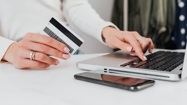 Mulher usando cartão de crédito para compras online Uma mulher, usando um anel de casamento e esmalte rosa claro, segura um cartão de crédito preto e branco na mão esquerda, enquanto usa a mão direita para digitar no teclado de um laptop prateado. Um smartphone preto está deitado ao lado do laptop na mesa branca. O fundo está embaçado, mas mostra roupas em um cabide.