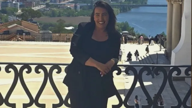 Mulher Sorridente em Miradouro Urbano Uma mulher sorridente com cabelo escuro pelos ombros, vestida com um blazer e blusa pretos, posa apoiada num corrimão de ferro forjado decorado. Ao fundo, vê-se um pátio amplo com algumas pessoas a caminhar, telhados de edifícios e um rio que corre sob uma ponte, num dia claro e ensolarado.