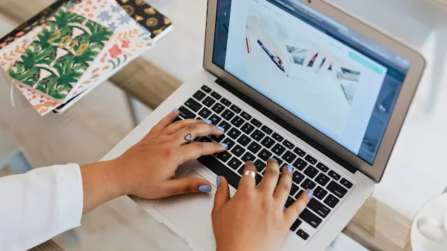Processo de criação de conteúdos e escrita num blog. Vista superior das mãos de uma pessoa com unhas pintadas de azul e uma tatuagem de coração no dedo, a digitar no teclado de um portátil MacBook Air. O ecrã mostra uma imagem de alguém a desenhar ou a escrever. Ao lado, sobre a mesa de vidro, destacam-se cadernos com capas coloridas e padrões florais.