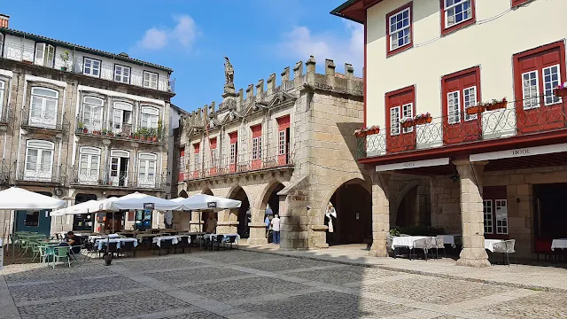 Praça da Oliveira no Centro Histórico de Guimarães Vista da Praça da Oliveira, em Guimarães, Portugal, num dia ensolarado. Ao centro, destaca-se o Antigo Paço do Concelho, um edifício medieval de pedra com arcos góticos no rés-do-chão e ameias no topo. À esquerda e à direita, encontram-se edifícios históricos tradicionais com varandas de ferro forjado e portadas coloridas. No piso térreo, esplanadas de restaurantes com mesas e guarda-sóis brancos ocupam o chão de calçada portuguesa, com algumas pessoas a caminhar e sentadas. O céu está azul com nuvens ligeiras.