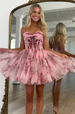 Floral Print Mini Dress with Corset Bodice A smiling young woman with blonde hair poses in front of a mirror wearing a pink, floral-print mini dress with thin straps, a structured corset-style bodice, and a voluminous, layered tulle skirt.