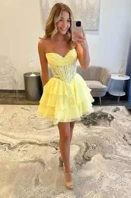 Shine at Your Next Event: The Perfect Yellow Homecoming Dress A young woman with wavy blonde hair smiles while posing for a mirror selfie, holding a silver smartphone. She is wearing a strapless, short yellow dress featuring a structured corset bodice and a ruffled, layered skirt. She completes the look with clear high-heeled sandals. The reflection shows a dress boutique with a grey and white abstract-patterned rug, a transparent acrylic chair, a grey tufted sofa, and small side tables with lamps. The lighting is soft and bright.