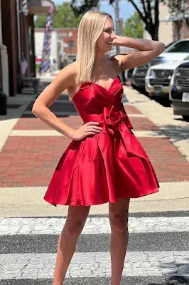 A smiling blonde young woman posing on a city crosswalk, wearing a short, red strapless satin homecoming dress featuring a large bow on the waist and a flared skirt. Elegant Red Strapless Homecoming Dress with Bow Detail